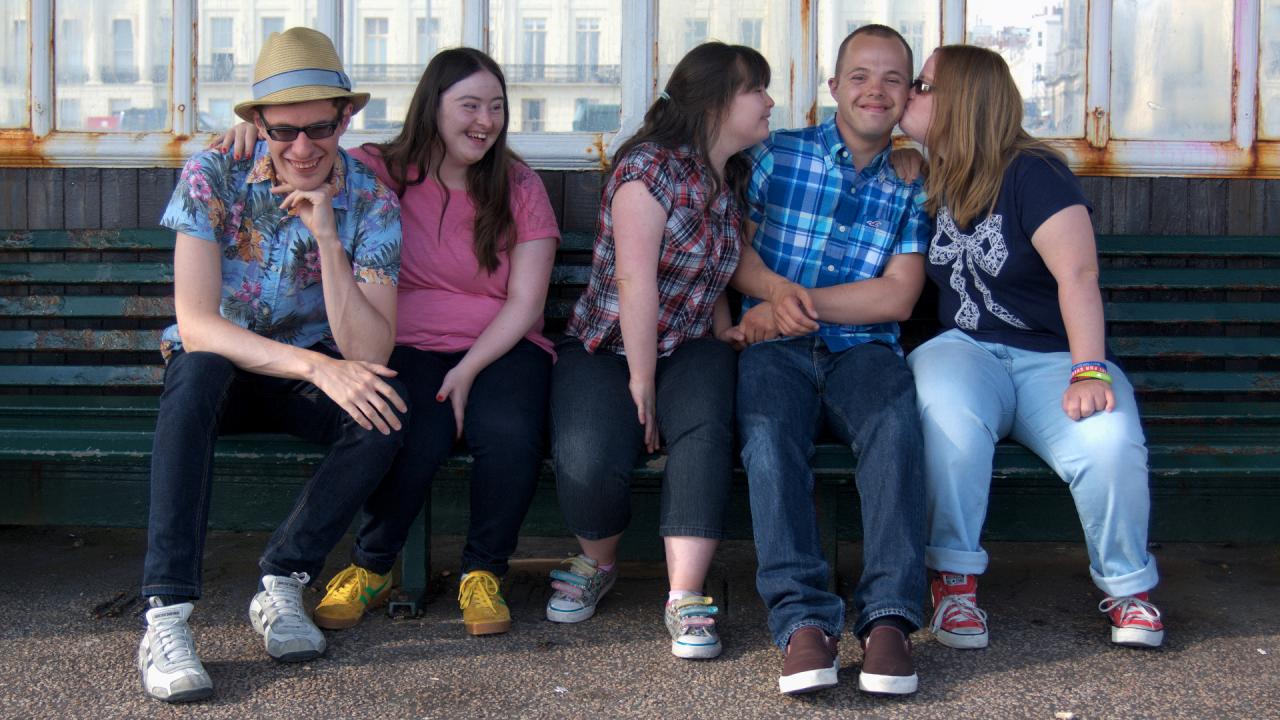 Lewis, Hilly, Megan, Sam and Lucy on a bench on Brighton promanade. Lucy is kissing Sam on the cheek.